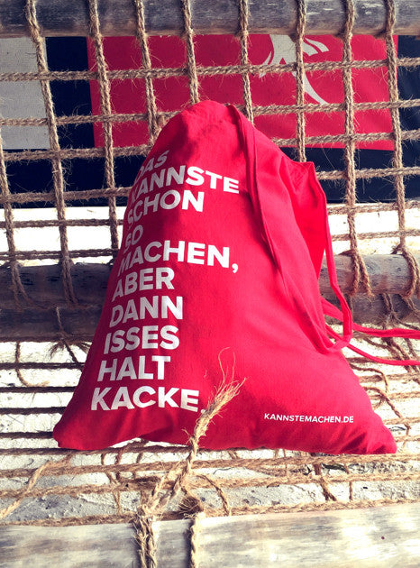 Der rote Jutebeutel, zugezogen auf einer Holzbank vor einem Fischernetz. Der Spruch steht in weißer Schrift darauf.