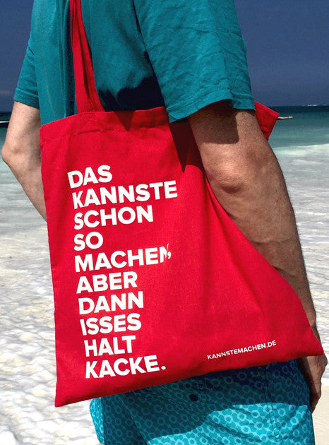 Der rote Jutebeutel über der Schulter getragen, am Strand. Der Spruch steht in weißer Schrift auf der Vorderseite.