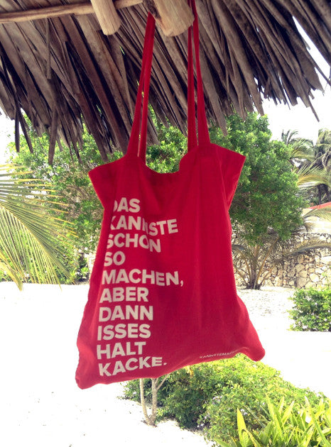 Der rote Jutebeutel hängt unter einem Palmdach, dahinter Grün und Sand. Auch hier trägt er den Spruch in weißer Schrift.
